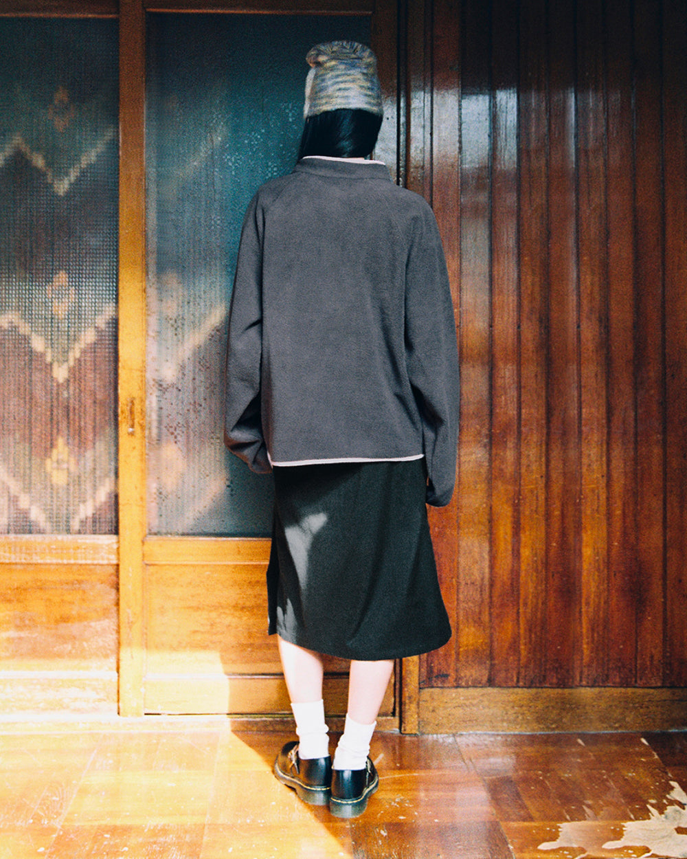A person stands indoors facing a wooden door, wearing the Sundae School CHARCOAL BUTTERFLY FLEECE PULLOVER with tteokbokki toggles, paired with a dark skirt, white socks, black shoes, and a knit beanie. Sunlight shines on the wooden floor.