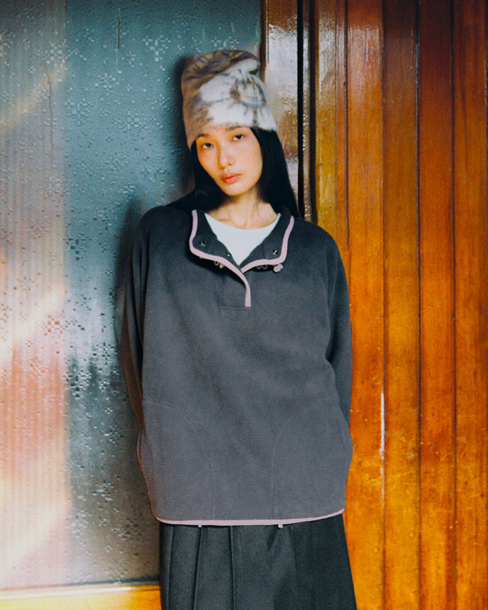A person wearing the Sundae School CHARCOAL BUTTERFLY FLEECE PULLOVER—a gray mock-neck polar fleece accented with pink trim and hand-tied flower knots—paired with a tie-dye beanie and a black skirt, stands indoors against a wooden wall and frosted glass, looking at the camera with a neutral expression.