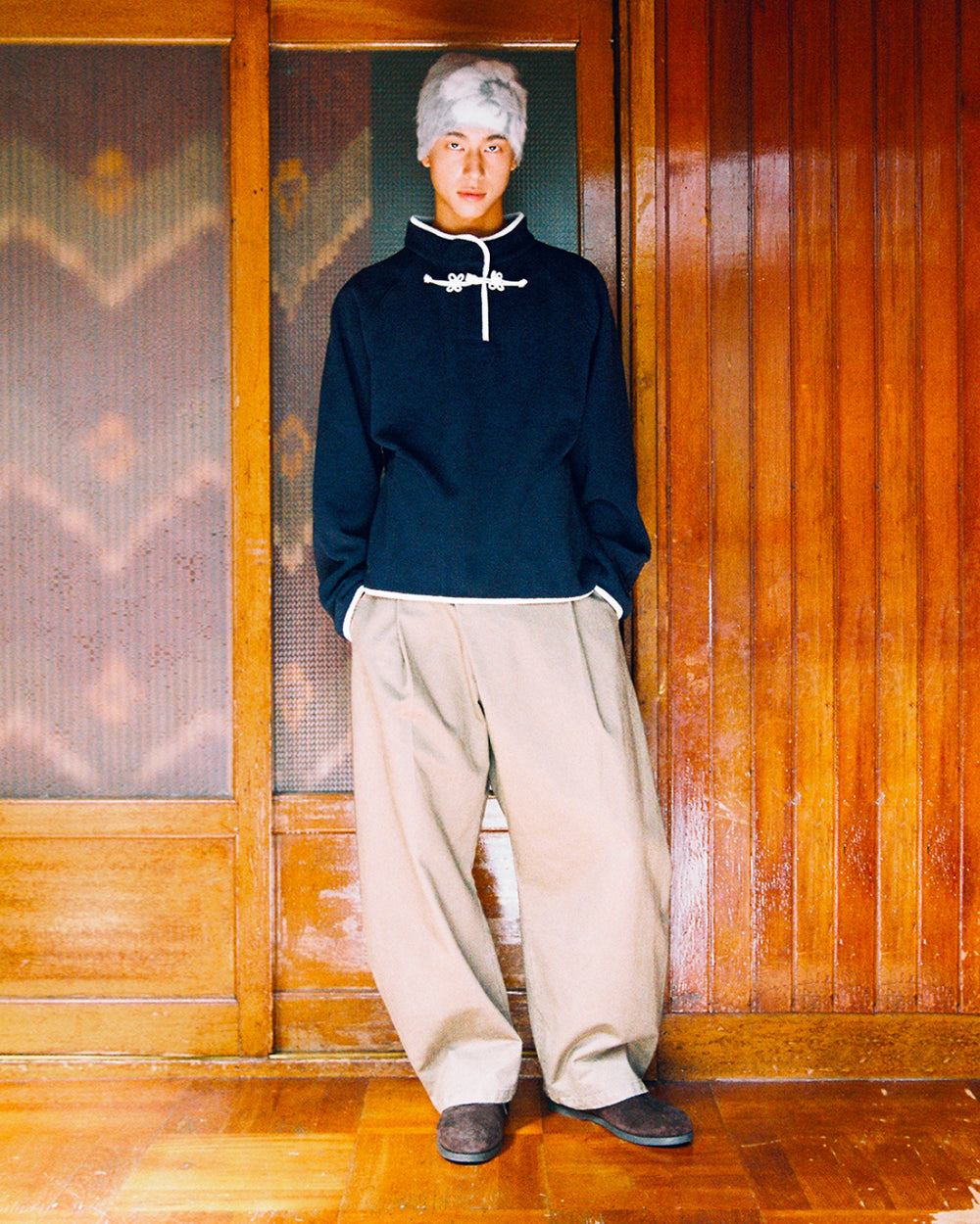 A person with short gray hair wears the Sundae School NAVY BUTTERFLY FLEECE PULLOVER and baggy beige pants, standing indoors against wooden sliding doors.