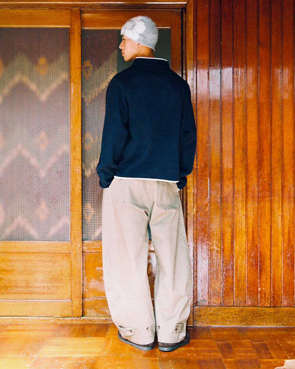 A person wearing a gray beanie and loose beige pants with flower knot toggles stands with their back to the camera in front of wooden sliding doors, dressed in the Sundae School NAVY BUTTERFLY FLEECE PULLOVER.