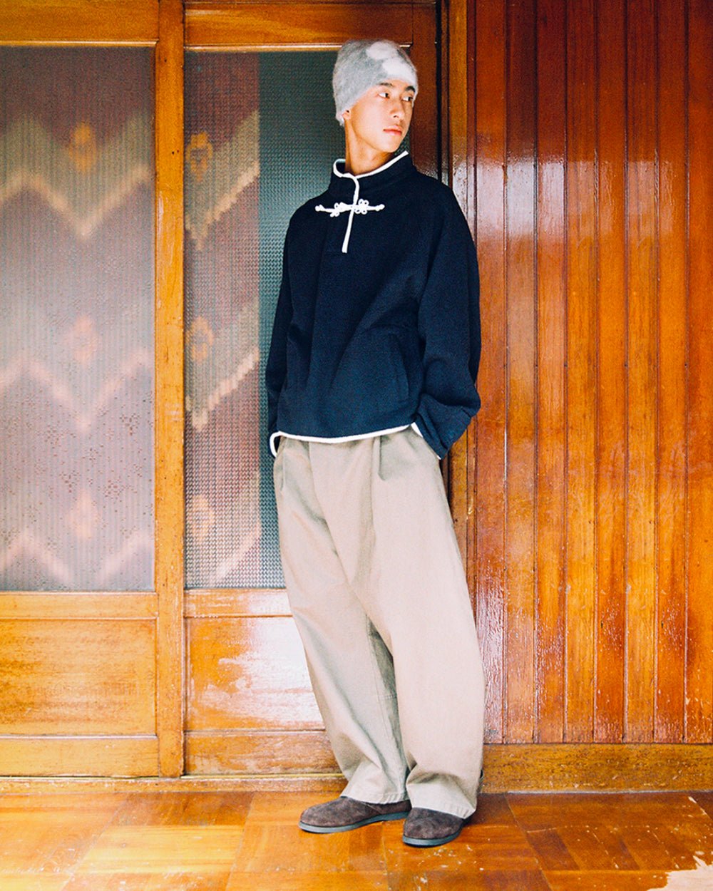 A person in a gray beanie, Sundae School’s NAVY BUTTERFLY FLEECE PULLOVER, wide khaki pants, and dark shoes stands indoors against wooden paneled walls and a frosted glass door, looking to the side with hands in their pockets.