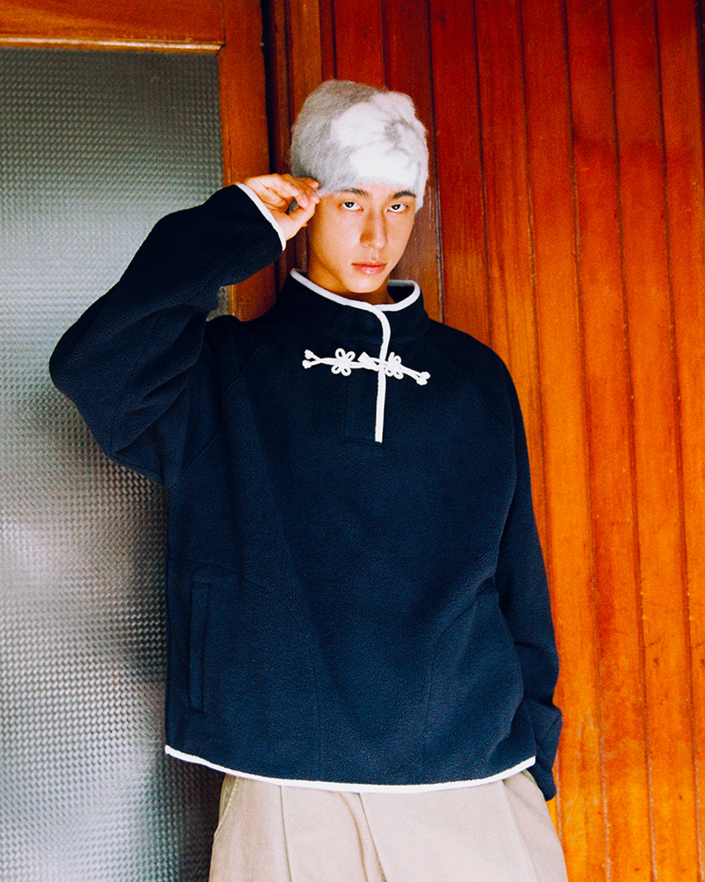 A person in the Sundae School NAVY BUTTERFLY FLEECE PULLOVER stands indoors, raising one hand by their head in front of a wooden wall and a frosted glass door. Their fluffy light gray hat completes the cozy look.