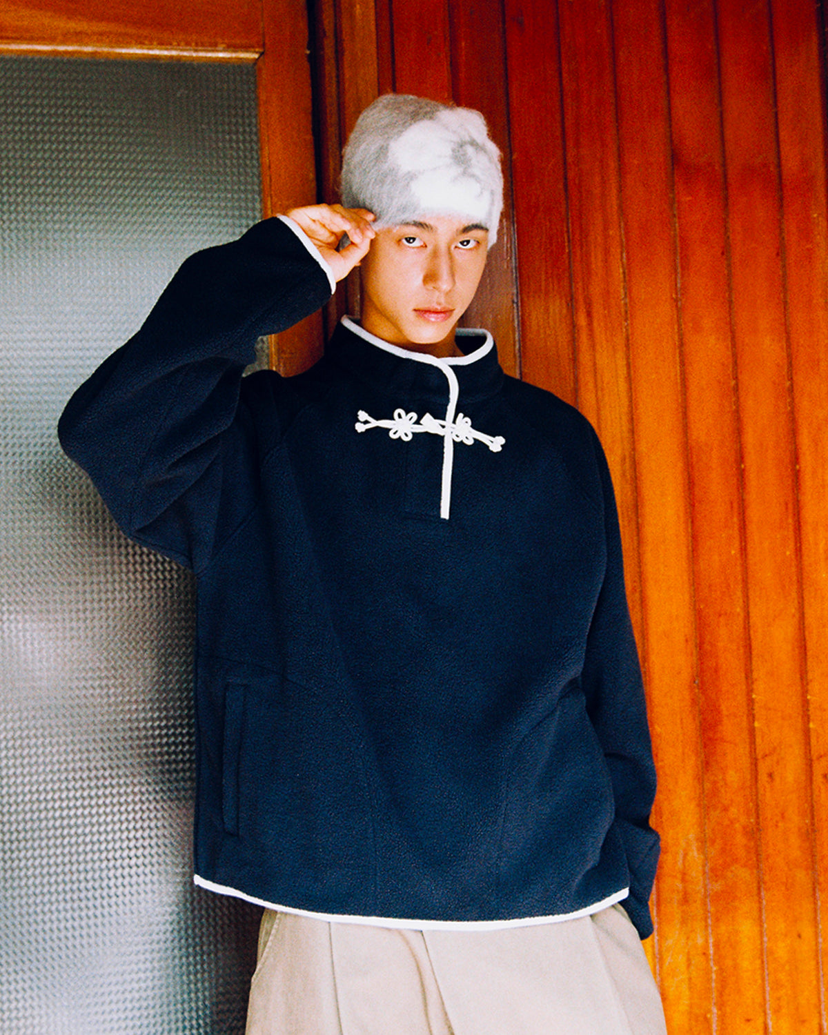 A person in the Sundae School NAVY BUTTERFLY FLEECE PULLOVER stands indoors, raising one hand by their head in front of a wooden wall and a frosted glass door. Their fluffy light gray hat completes the cozy look.