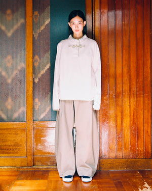 A person stands indoors against a wooden wall, wearing the Sundae School TAUPE BUTTERFLY FLEECE PULLOVER—a loose, light-colored mock-neck fleece—paired with wide beige pants. Their hair is neatly parted, and they face the camera calmly with hands by their sides.