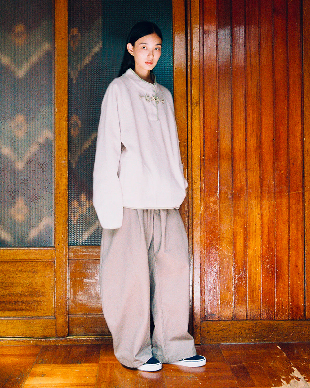 A person stands indoors against wooden paneling, wearing the Sundae School TAUPE BUTTERFLY FLEECE PULLOVER—a loose, light-colored long-sleeve top—paired with wide beige pants and black slip-on shoes, looking at the camera with a neutral expression.
