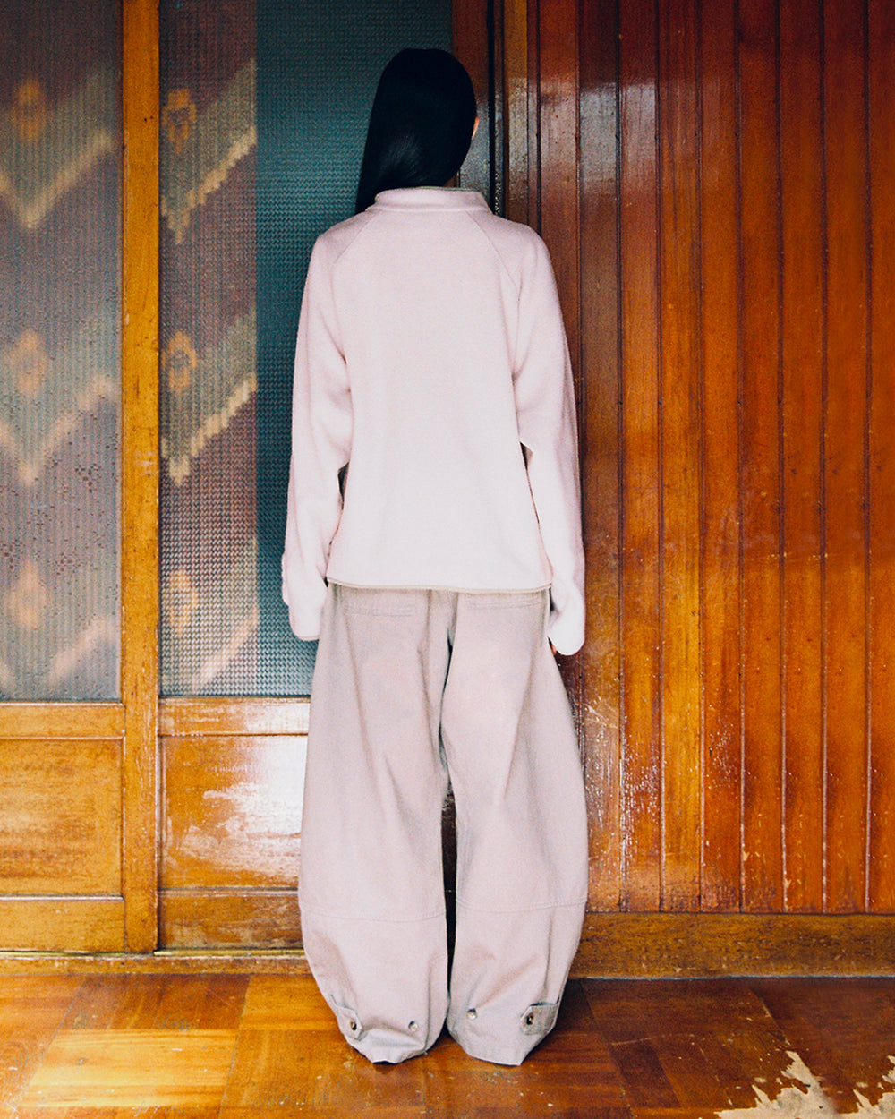 A person with long dark hair faces away, wearing the Sundae School TAUPE BUTTERFLY FLEECE PULLOVER and very baggy matching pants, standing in front of wooden doors on a polished wood floor.