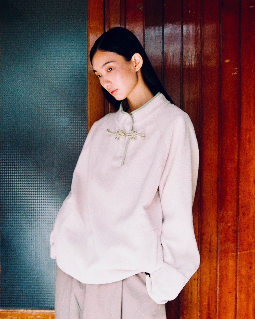 A person with long dark hair stands against a wooden wall, gazing slightly down and to the side with hands in their pockets, wearing the Sundae School TAUPE BUTTERFLY FLEECE PULLOVER—a pale, loose-fitting mock-neck style detailed with hand-tied flower knots.