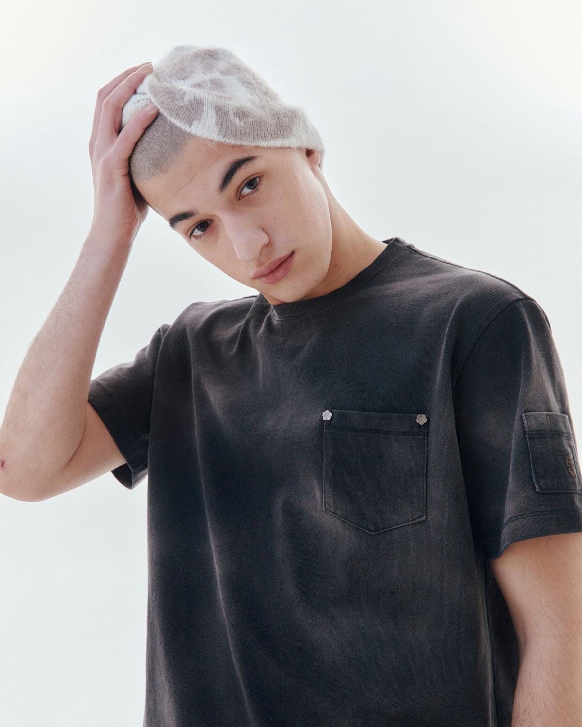 A young person in a black t-shirt and the Sundae School Ivory Ddul Beanie touches the top of their head, as if highlighting brain protection, while standing against a plain white background.