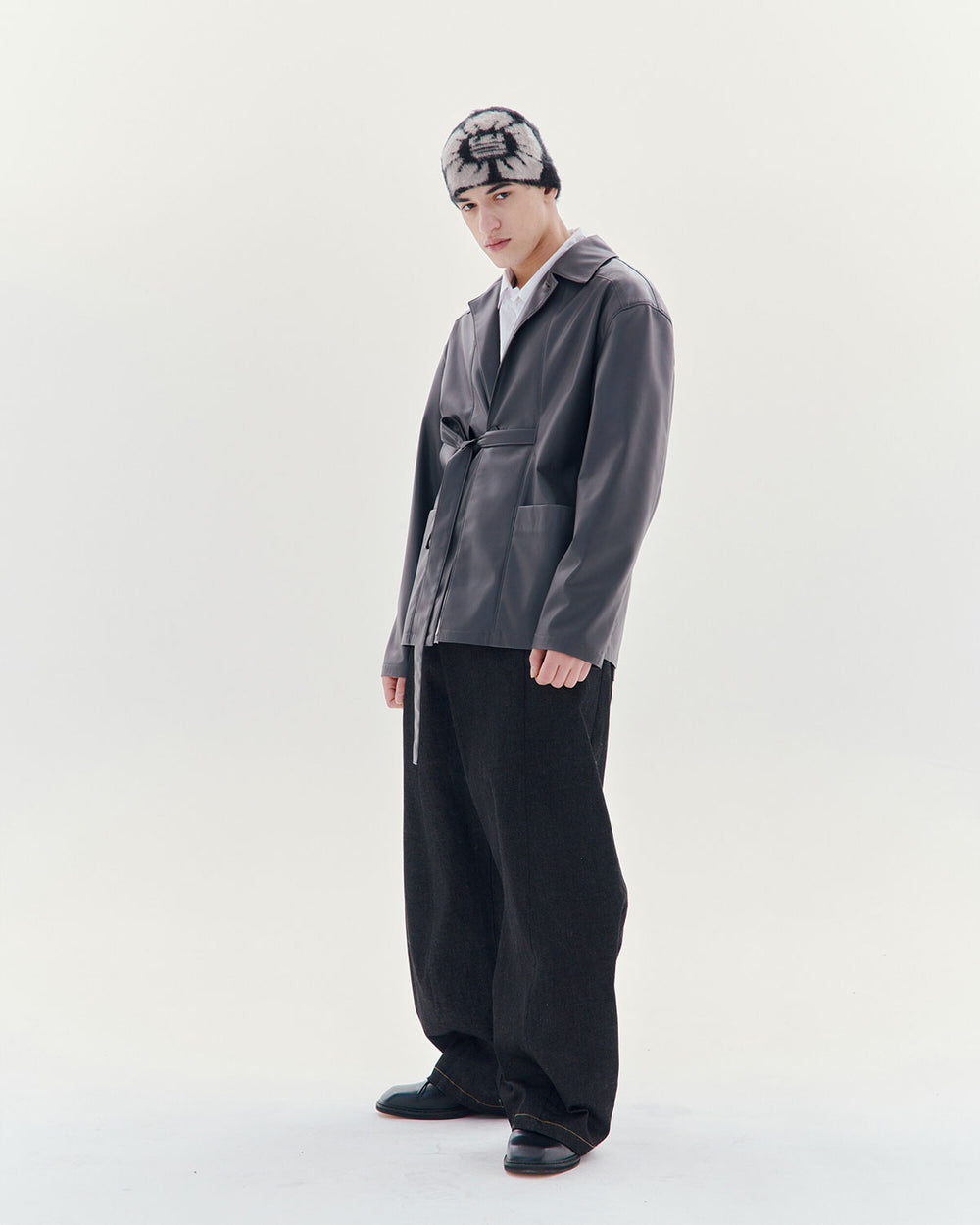 A person stands against a white background wearing a gray jacket, wide black pants, black shoes, and the Sundae School Black Ddul Beanie, featuring a black-and-white pattern, looking slightly down with a relaxed posture.