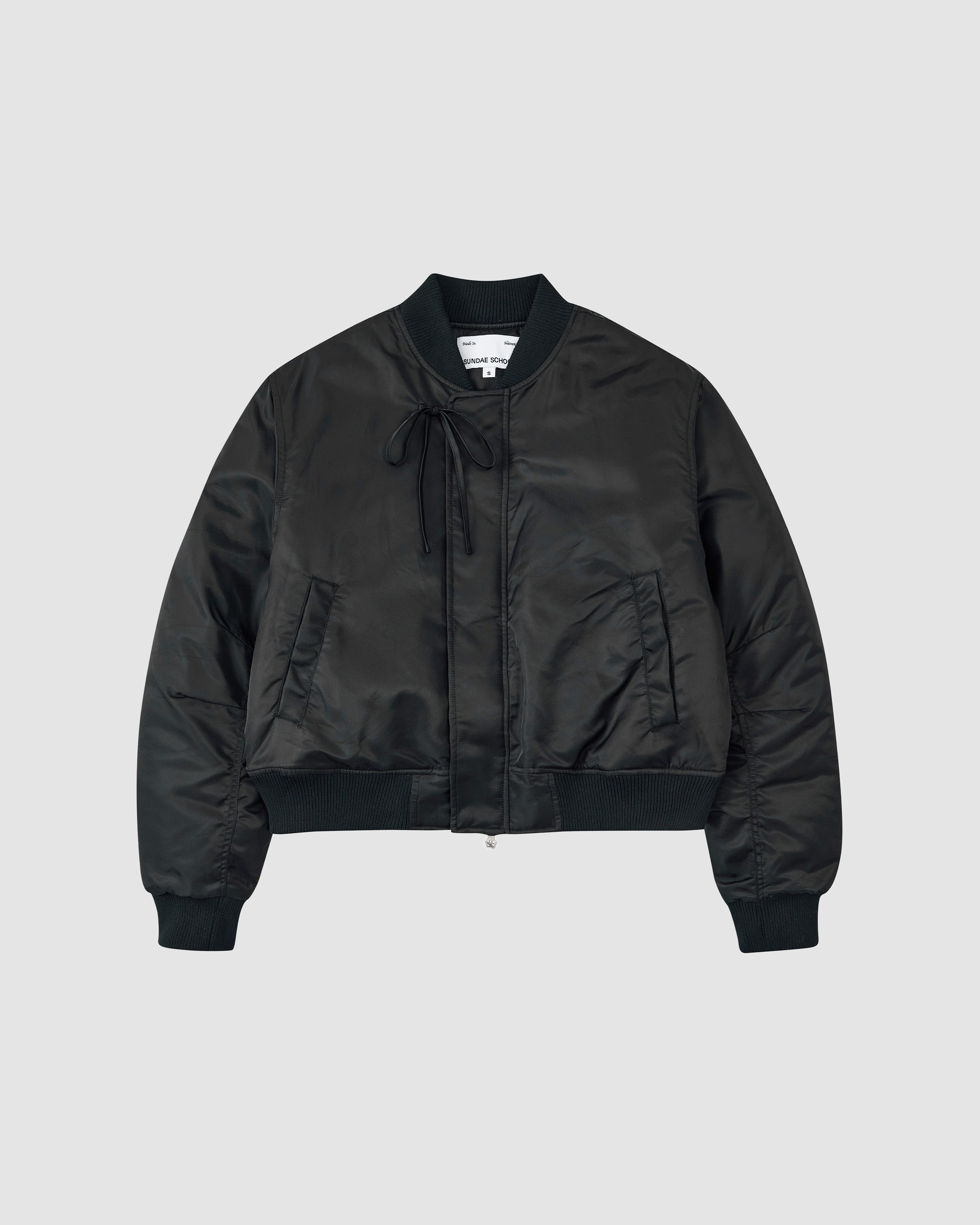 The BLACK FLOWER CHANNELED BOMBER by Sundae School features ribbed cuffs, collar, and hem, a front zip closure, side pockets, and a tie detail near the collar, shown against a plain light gray background.