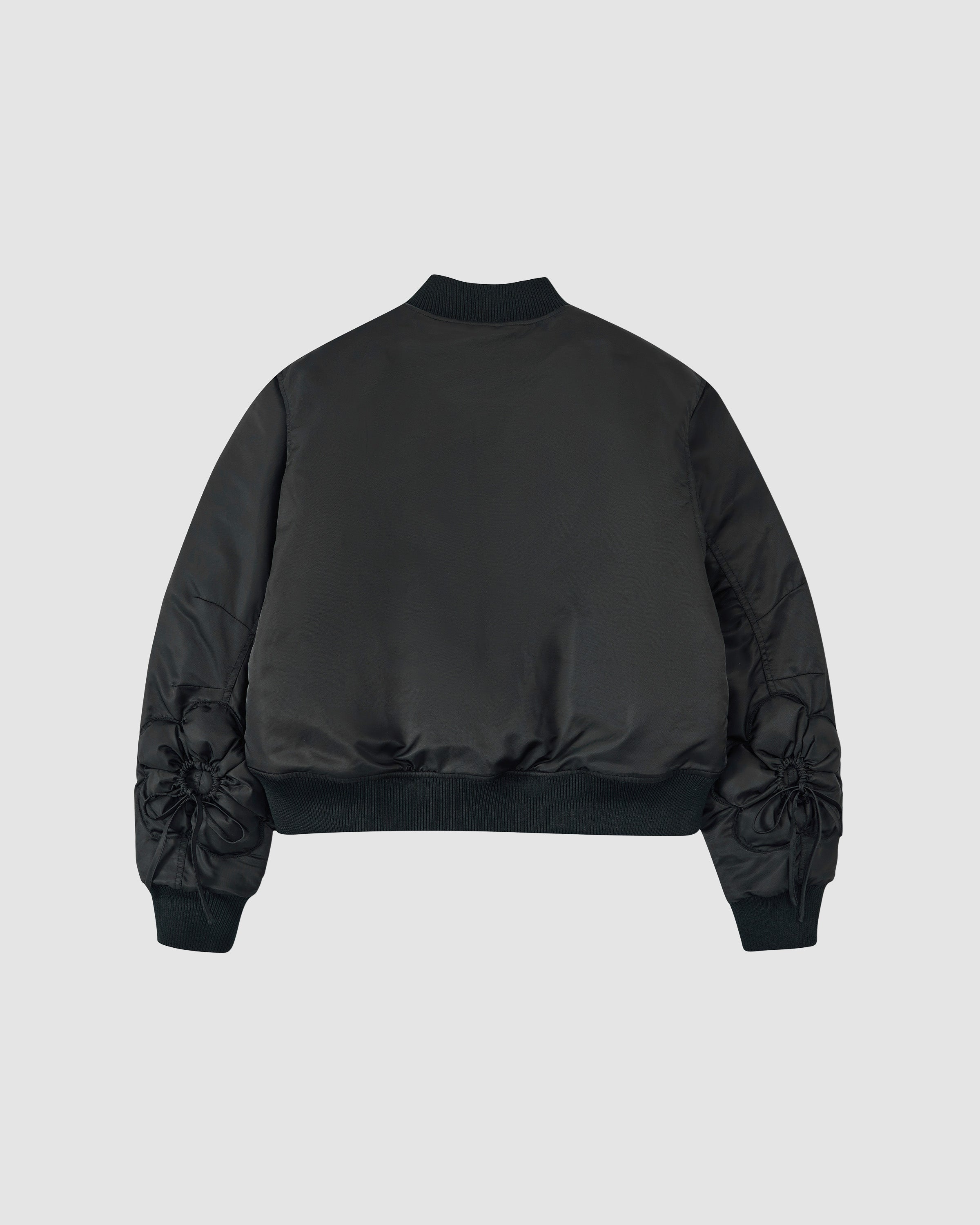 The BLACK FLOWER CHANNELED BOMBER by Sundae School is a black bomber jacket with a ribbed collar, cuffs, and hem. The plain back contrasts with sleeves featuring gathered, flower-like fabric details near the cuffs.