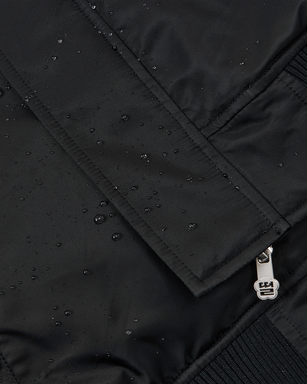 Close-up of the Sundae School BLACK FLOWER CHANNELED BOMBER shows water droplets highlighting its water-resistant fabric, along with a silver zipper and part of a pocket flap.