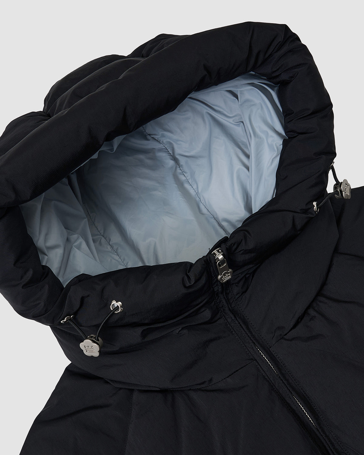 Close-up of the Sundae School BLACK SKY BLUE ORIGAMI PUFFER, a black hooded jacket with light gray inner lining, drawstring toggles, and a partially visible front zipper, laid flat on a plain light background.