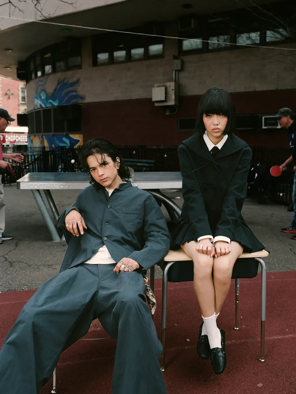 Two models pose in an urban outdoor setting. The model on the left wears SUNDAE SCHOOL STAND COLLAR UNIFORM JACKET and matching PLEATED UNIFORM PAPNTS over a white undershirt and the model on the right wears SUNDAE SCHOOL SAILOR COLLAR UNIFORM JACKET and APRON SKIRT.