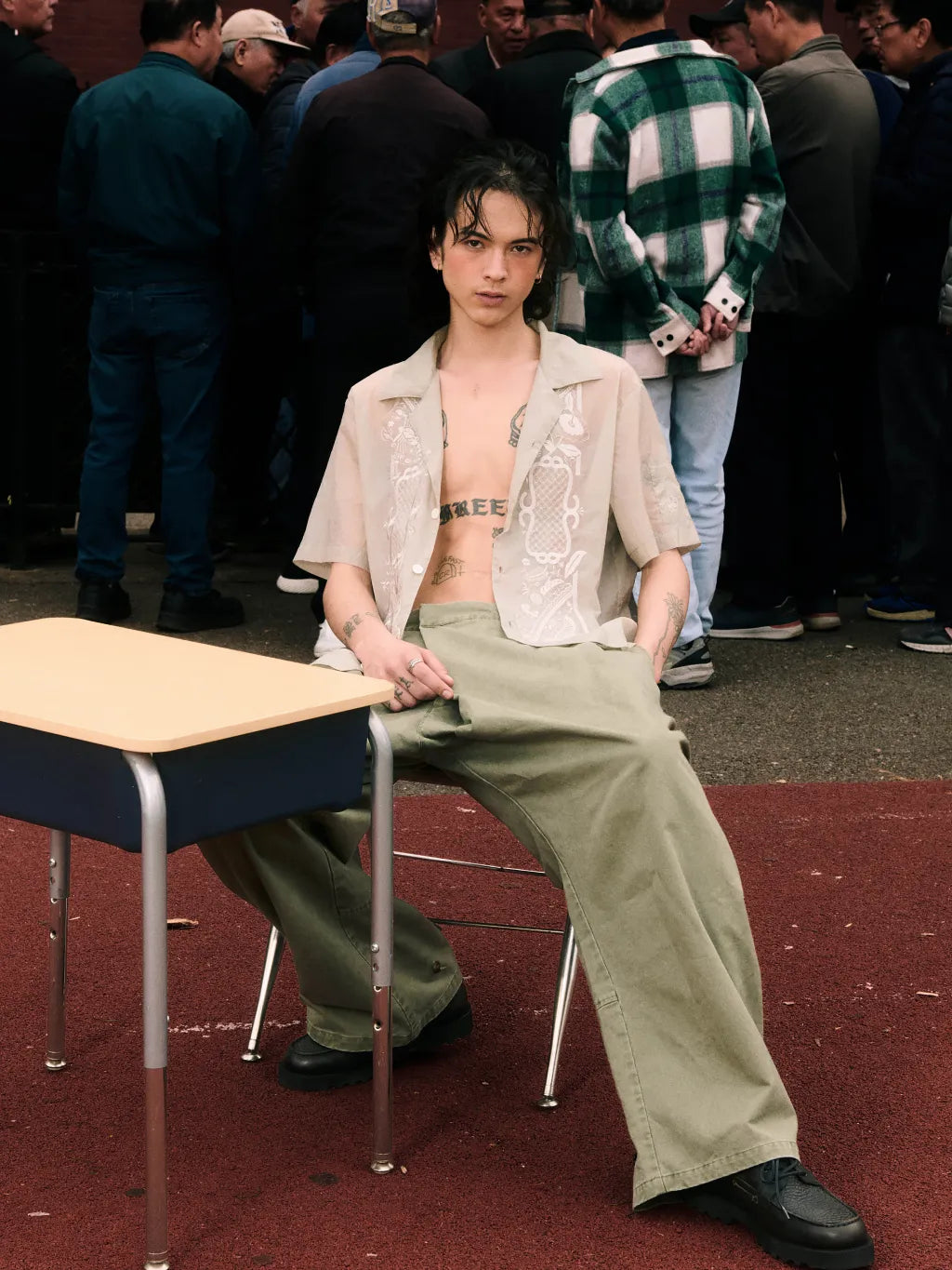 A person seated on a chair in front of a school desk, wearing  SUNDAE SCHOOL BARONG SHIRT over a bare torso with SUNDAE SCHOOL OLIVE HANBOK CHINO PANTS and black shoes. A crowd is visible in the background, and the scene appears to be an outdoor or event-like setting.