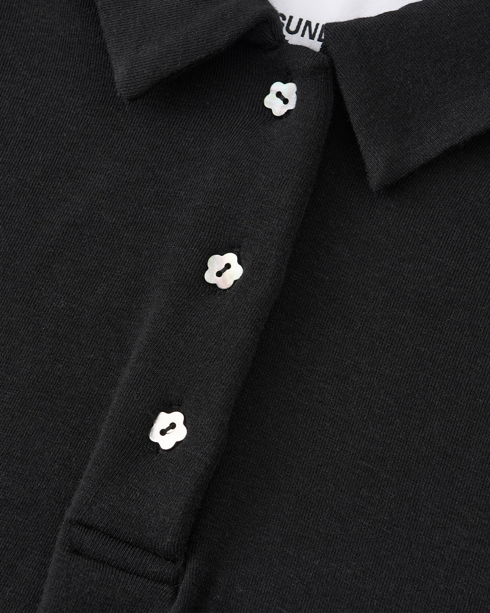 Close-up of the BLACK CINDY POLO DRESS by Sundae School, featuring a black collar and three iridescent, flower-shaped buttons on the placket—details that evoke classic sophistication. The edge of a white shirt label is partially visible inside the collar.