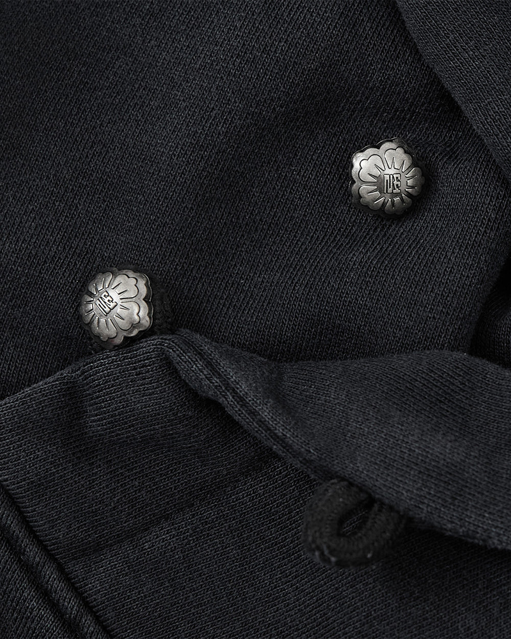 Close-up of two silver floral metal buttons on dark fabric, with one button partly covered by a folded section of the material—an elegant detail that perfectly complements Sundae School’s FADE BLACK COCOON HOODIE.