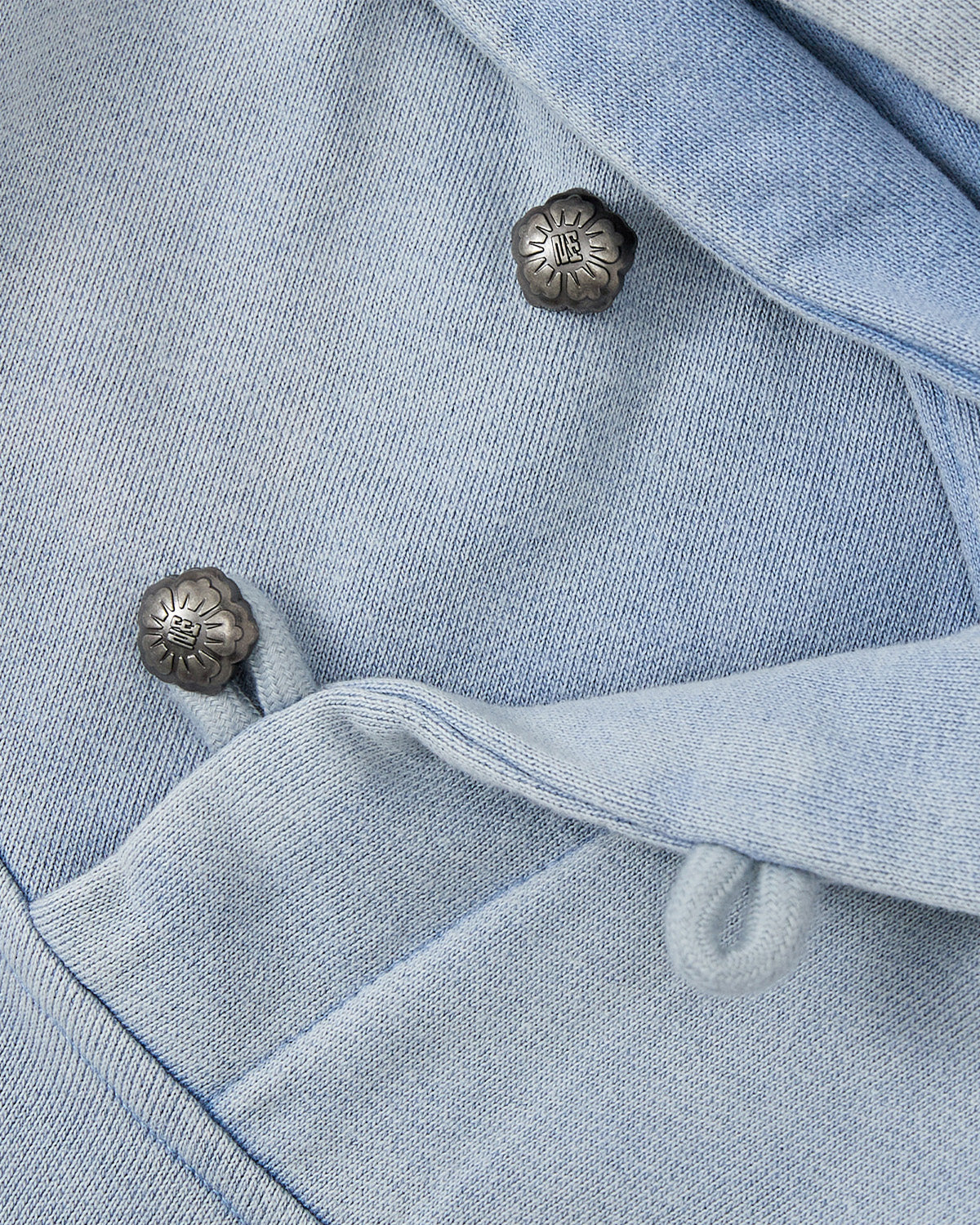 Close-up of SKY BLUE COCOON HOODIE by Sundae School, featuring soft light blue fabric with metal floral buttons and looped fabric fasteners, highlighting its winged silhouette and peret sleeves.
