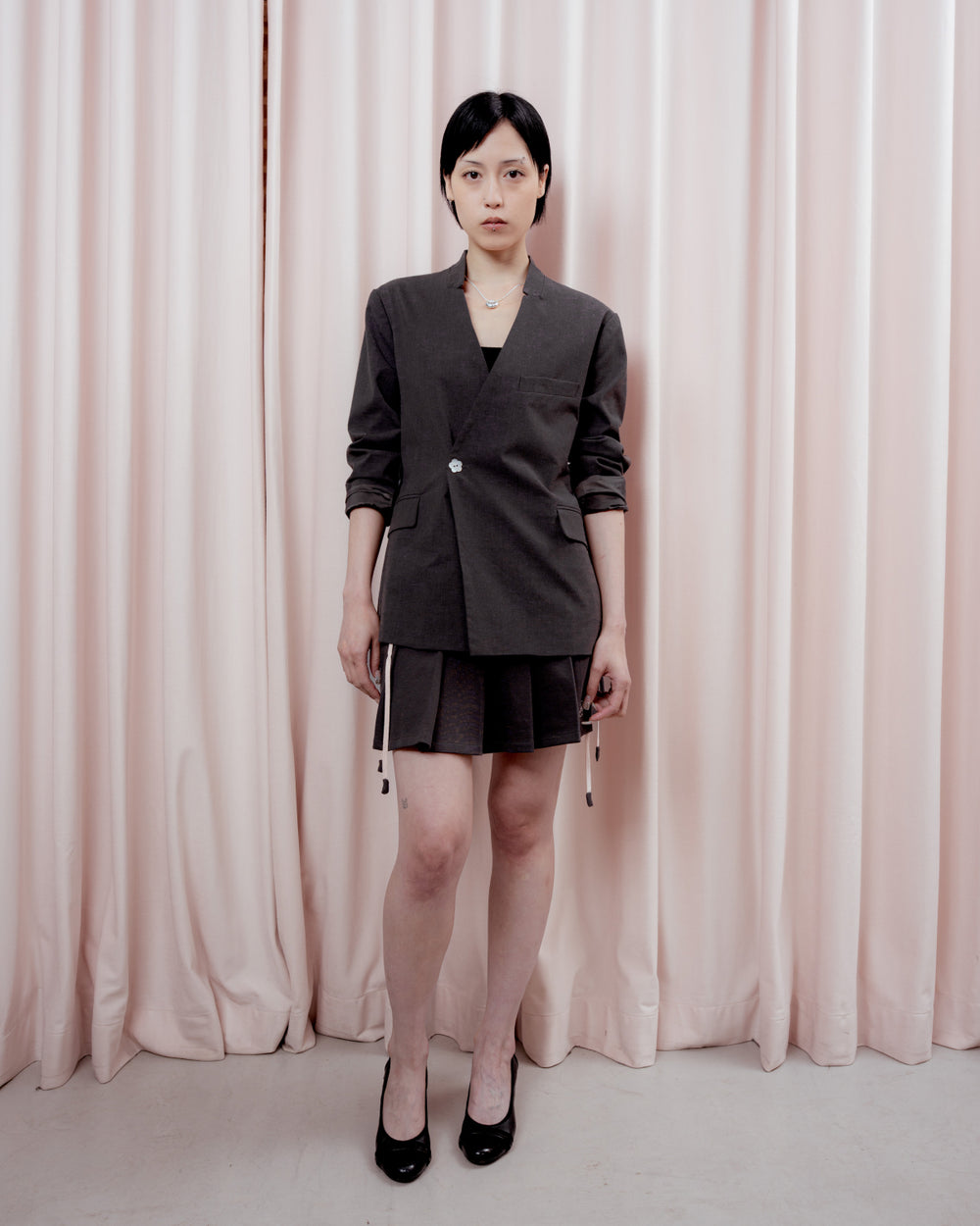 Full-length front view of a person with short black hair standing in front of a pink curtain backdrop. They are wearing a charcoal gray collarless blazer fastened with a single light-colored button, a matching pleated charcoal mini skirt with white drawstring ties hanging down, and black low heels.