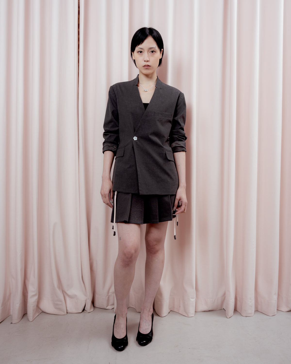 Full-length front view of a person with short black hair standing in front of a pink curtain backdrop. They are wearing a charcoal gray collarless blazer fastened with a single light-colored button, a matching pleated charcoal mini skirt with white drawstring ties hanging down, and black low heels.