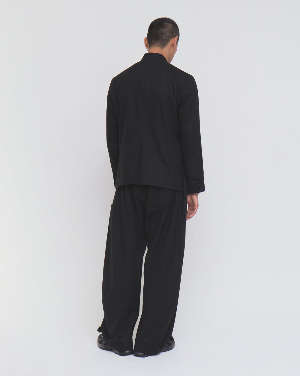 A person with short dark hair stands facing away, wearing Sundae School’s BLACK KOREAN AMERICAN BLAZER—a loose-fitting suit with jeogori-inspired paneling—paired with wide-legged pants and black shoes, set against a plain white background.