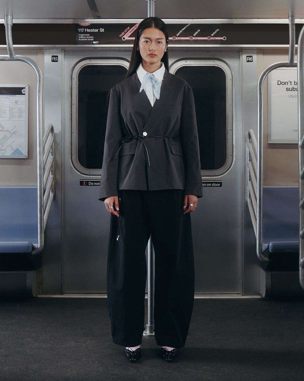 A woman stands in a subway car wearing the BLACK FLOWER NORIGAE LINEN TROUSERS from Sundae School, styled with a matching black suit and a white collared shirt. She faces forward between empty seats, with subway maps and signs visible behind her.