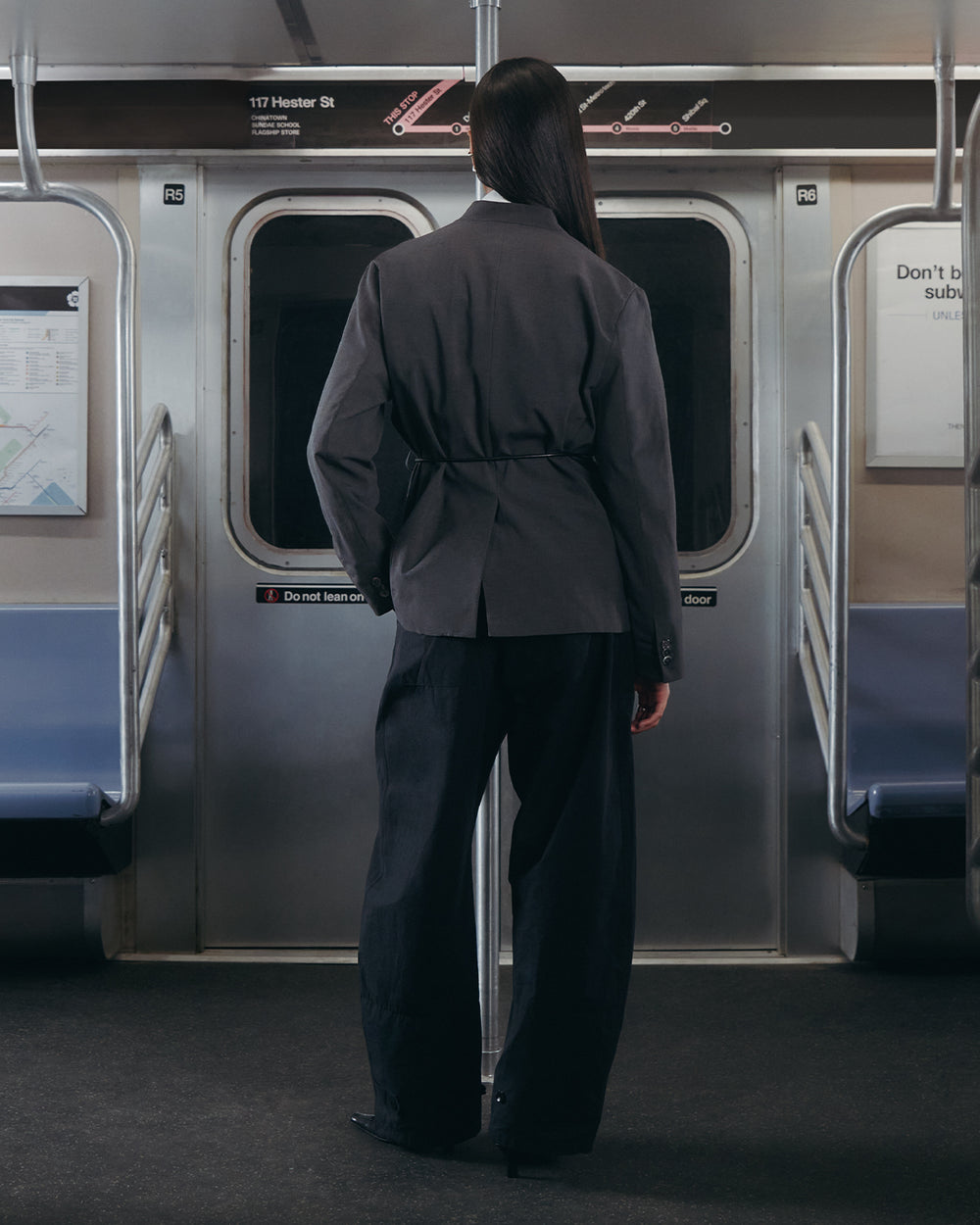 A person with long dark hair, wearing Sundae School's COLLARLESS TIE BLAZER and a vegan leather belt, stands facing the closed doors inside an empty subway train with maps on the walls.