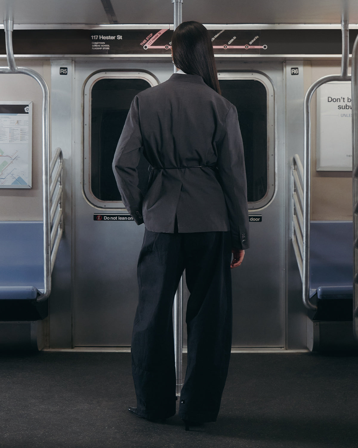 A person with long dark hair, dressed in Sundae School’s BLACK FLOWER NORIGAE LINEN TROUSERS, stands facing the subway doors inside a mostly empty subway car, with subway maps and signs visible on the walls.