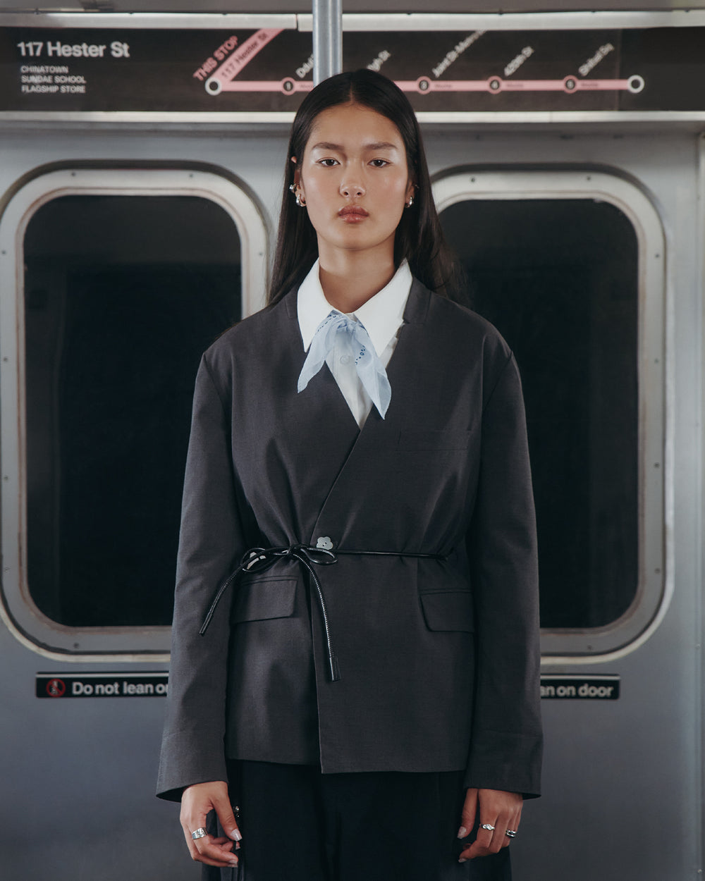 A woman with long, straight hair and a neutral expression stands in a subway car, wearing the Sundae School COLLARLESS TIE BLAZER with a light blue scarf and vegan leather belt. Behind her are a subway map and the 117 Hester St sign.