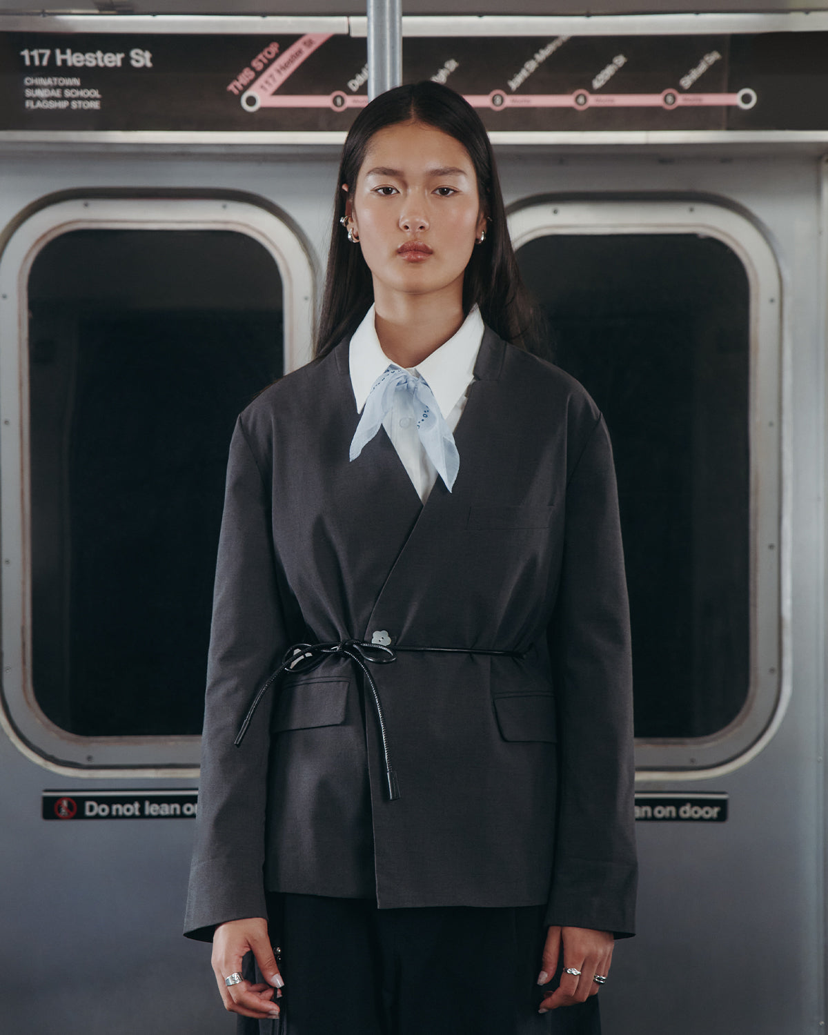 A woman with long, straight hair and a neutral expression stands in a subway car, wearing the Sundae School COLLARLESS TIE BLAZER with a light blue scarf and vegan leather belt. Behind her are a subway map and the 117 Hester St sign.