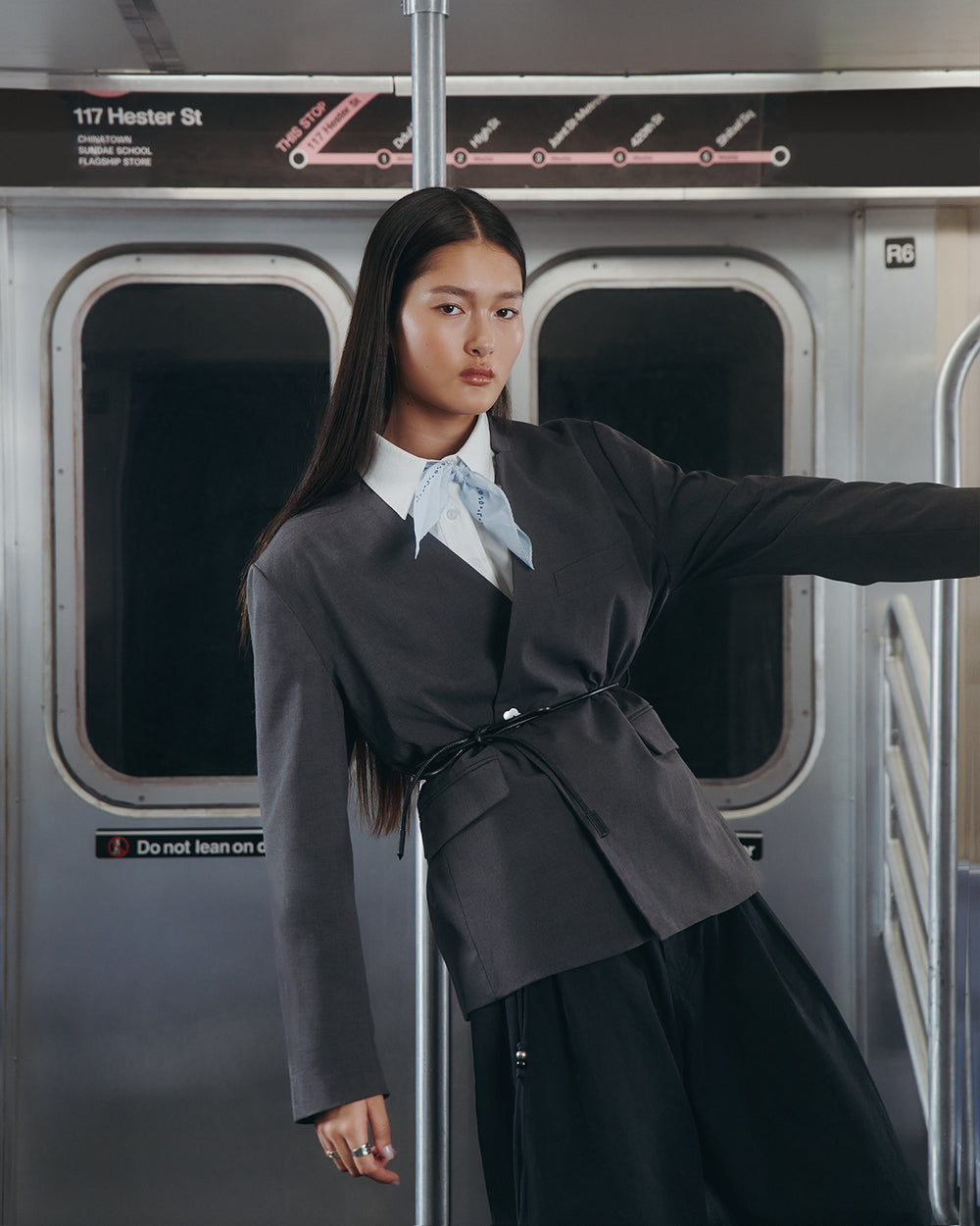 A woman in the Sundae School COLLARLESS TIE BLAZER, paired with a light blue scarf and black pants, poses confidently inside a subway car. A station sign reading 117 Hester St is visible above the door behind her.