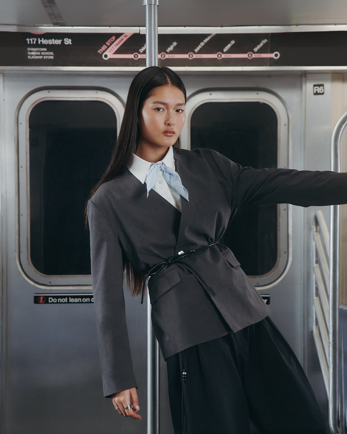 A woman in the Sundae School COLLARLESS TIE BLAZER, paired with a light blue scarf and black pants, poses confidently inside a subway car. A station sign reading 117 Hester St is visible above the door behind her.