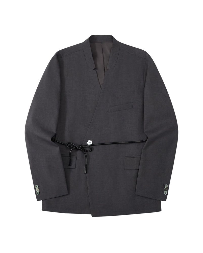 The COLLARLESS TIE BLAZER by Sundae School is a dark gray blazer with an unstructured drape and a shawl collar, designed without buttons. It features a single chest pocket, two front flap pockets, and comes with a thin vegan leather belt that ties at the waist.