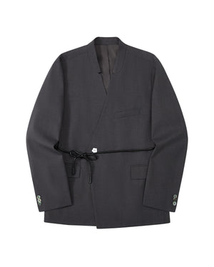 The COLLARLESS TIE BLAZER by Sundae School is a dark gray blazer with an unstructured drape and a shawl collar, designed without buttons. It features a single chest pocket, two front flap pockets, and comes with a thin vegan leather belt that ties at the waist.
