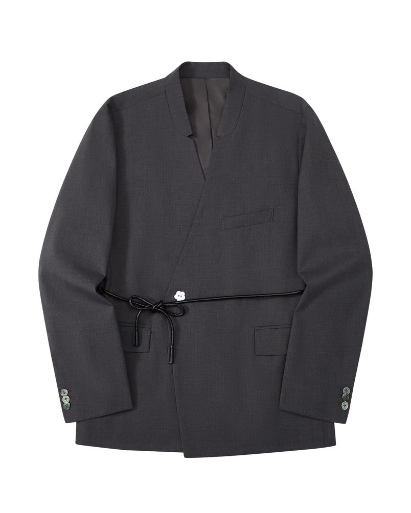 The COLLARLESS TIE BLAZER by Sundae School is a dark gray blazer with an unstructured drape and a shawl collar, designed without buttons. It features a single chest pocket, two front flap pockets, and comes with a thin vegan leather belt that ties at the waist.