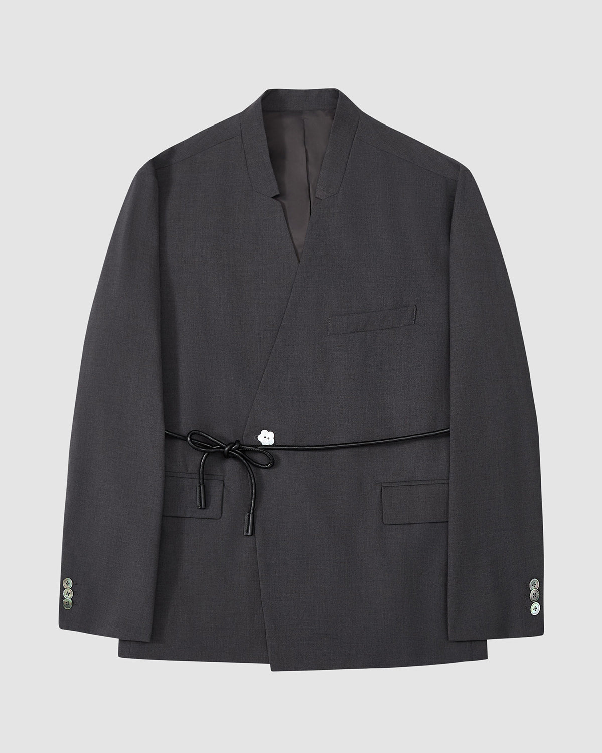 The Sundae School COLLARLESS TIE BLAZER is a dark gray, collarless summer wool blazer with a wrap front, chest pocket, two flap pockets, buttoned cuffs, and a vegan leather waist belt, shown on a plain light gray background.