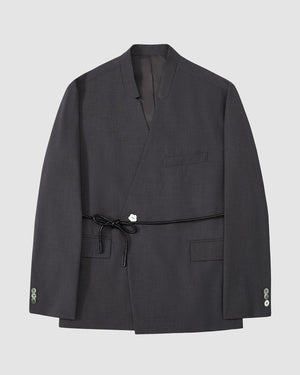 The Sundae School COLLARLESS TIE BLAZER is a dark gray, collarless summer wool blazer with a wrap front, chest pocket, two flap pockets, buttoned cuffs, and a vegan leather waist belt, shown on a plain light gray background.