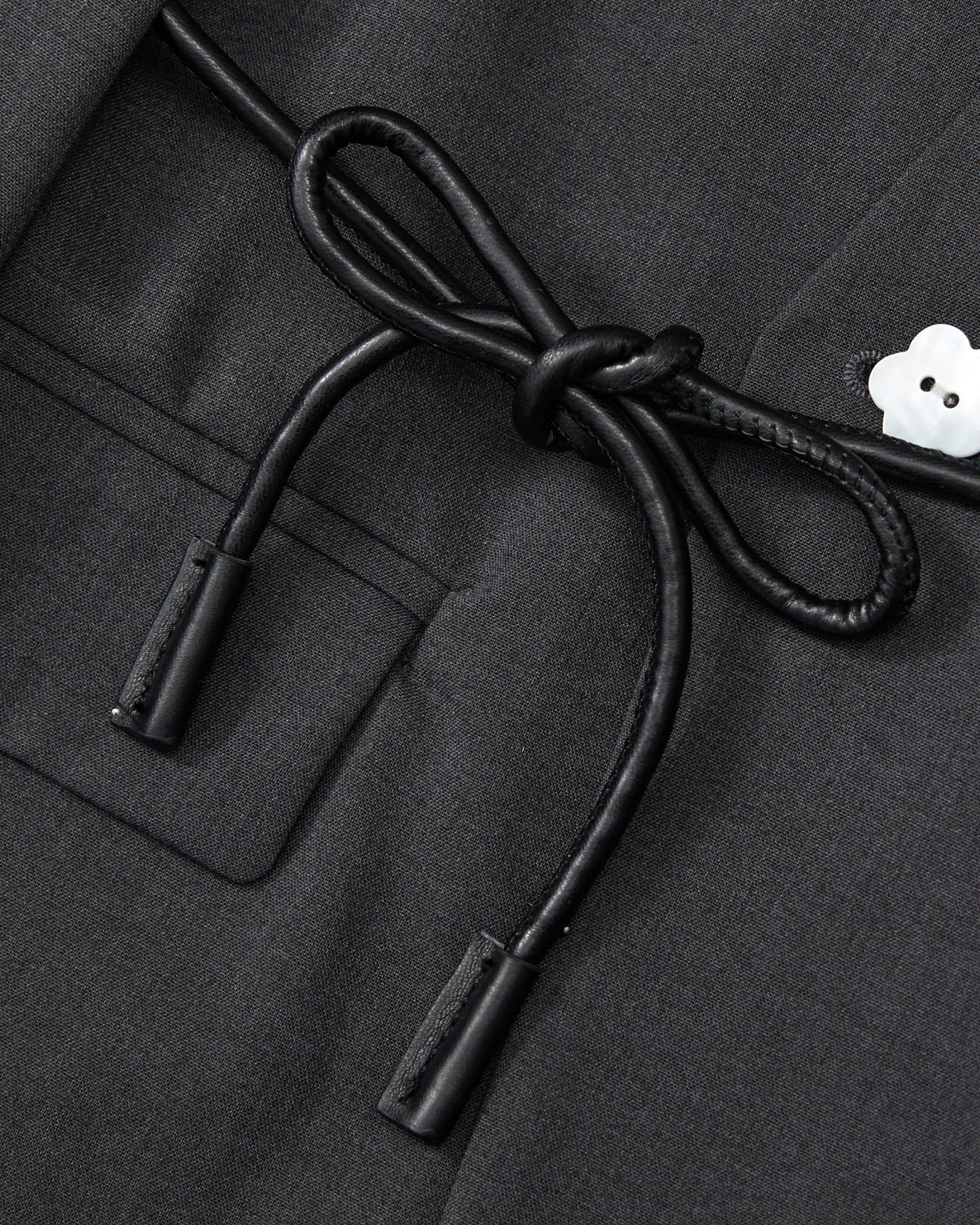 Close-up of the COLLARLESS TIE BLAZER by Sundae School in black, showcasing its unstructured drape and pocket, accented with a black vegan leather cord belt tied in a bow and finished with a small white flower-shaped button on the right.