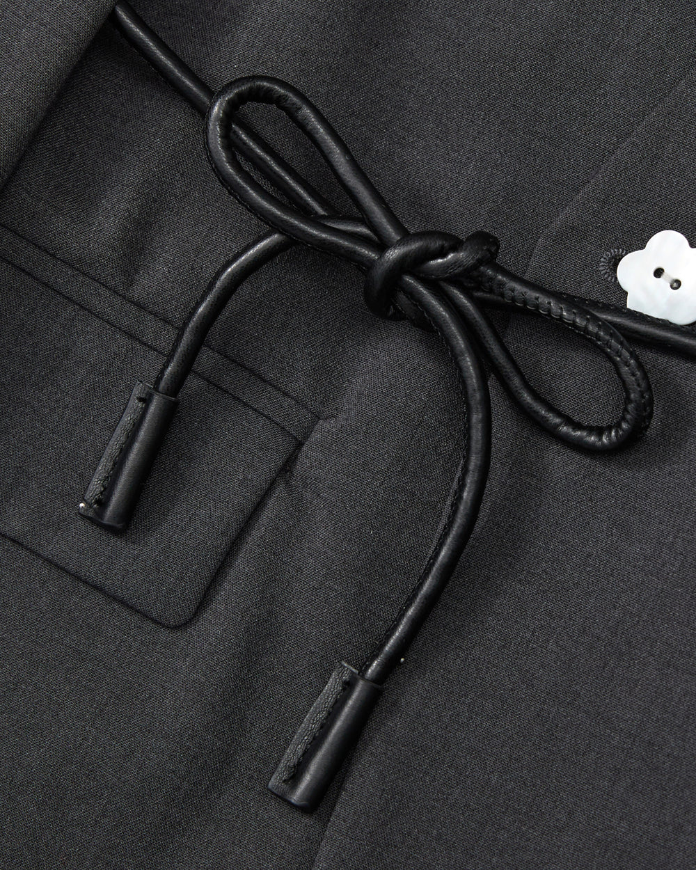 Close-up of the COLLARLESS TIE BLAZER by Sundae School in black, showcasing its unstructured drape and pocket, accented with a black vegan leather cord belt tied in a bow and finished with a small white flower-shaped button on the right.