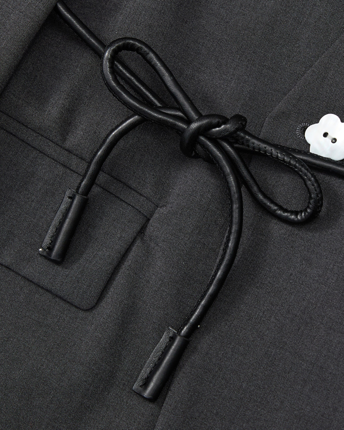 Close-up of the COLLARLESS TIE BLAZER by Sundae School in black, showcasing its unstructured drape and pocket, accented with a black vegan leather cord belt tied in a bow and finished with a small white flower-shaped button on the right.