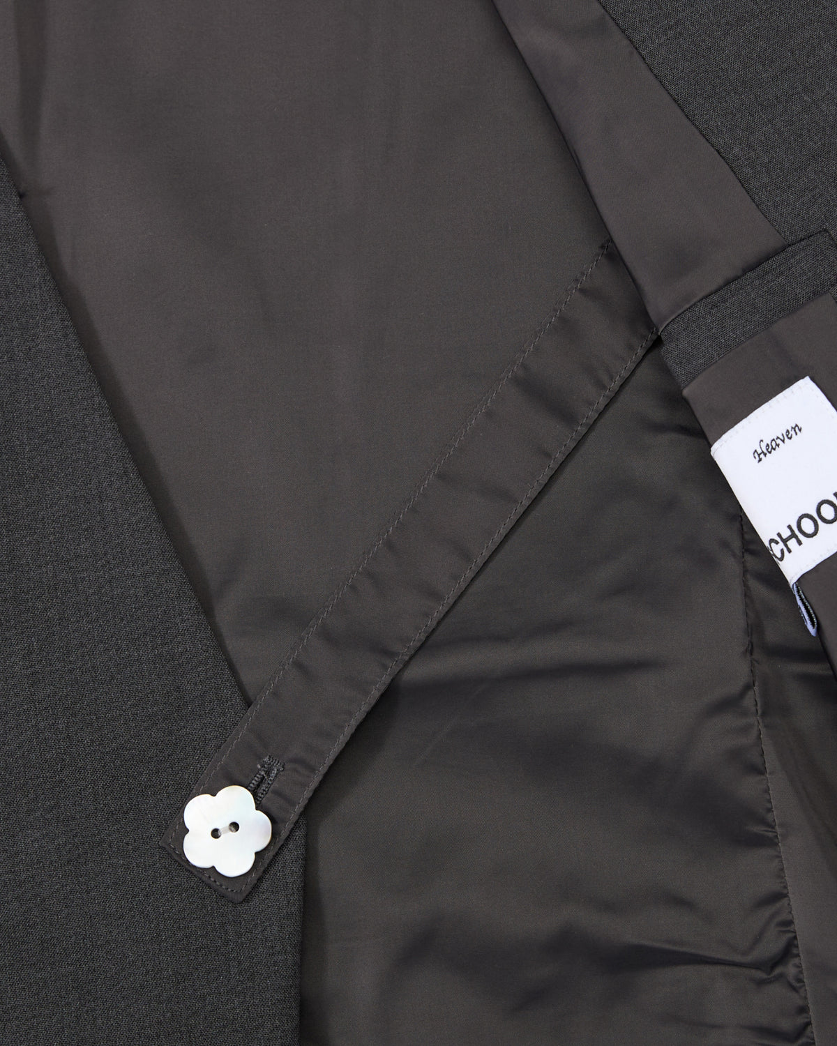 A close-up of the inside lining of Sundae School’s COLLARLESS TIE BLAZER in dark gray, featuring a buttoned strap with a white flower-shaped button and a partially visible Sundae School label, highlighting the blazer’s unstructured drape.