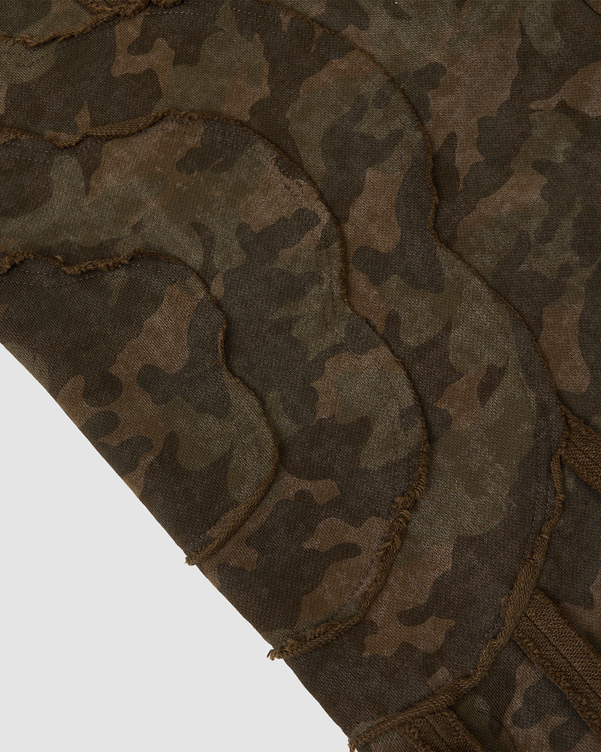 Close-up of Sundae School's CAMO MOCHI HAZE PANELED SWEATPANTS, crafted from 100% cotton in a green and brown camouflage pattern with curved, raw-edged stitched lines. Set against a light gray background.