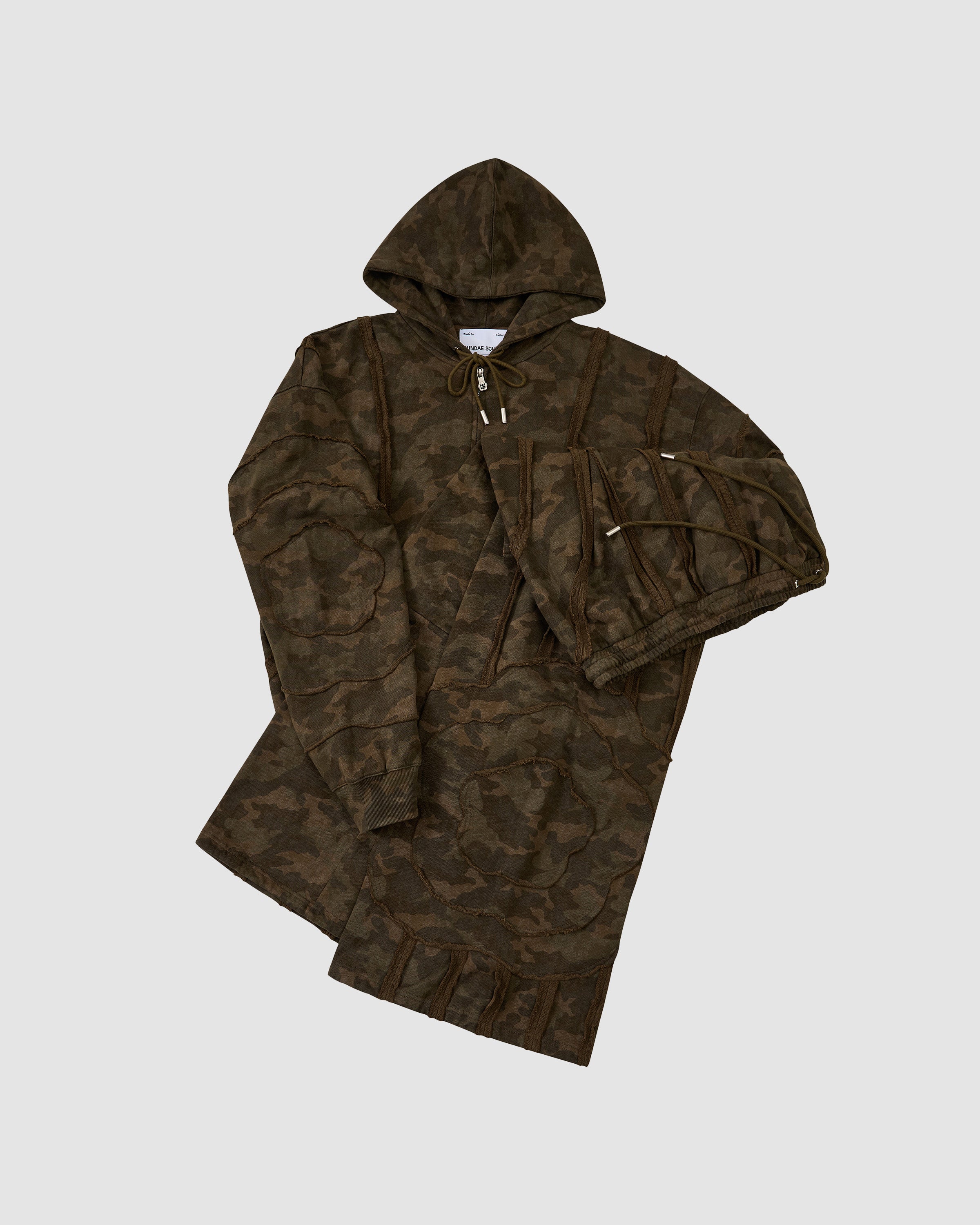 The Sundae School CAMO MOCHI HAZE PANELED ZIP UP HOODIE, featuring a camouflage pattern and raw seam panels in 100% cotton, is displayed laid flat on a light gray background.
