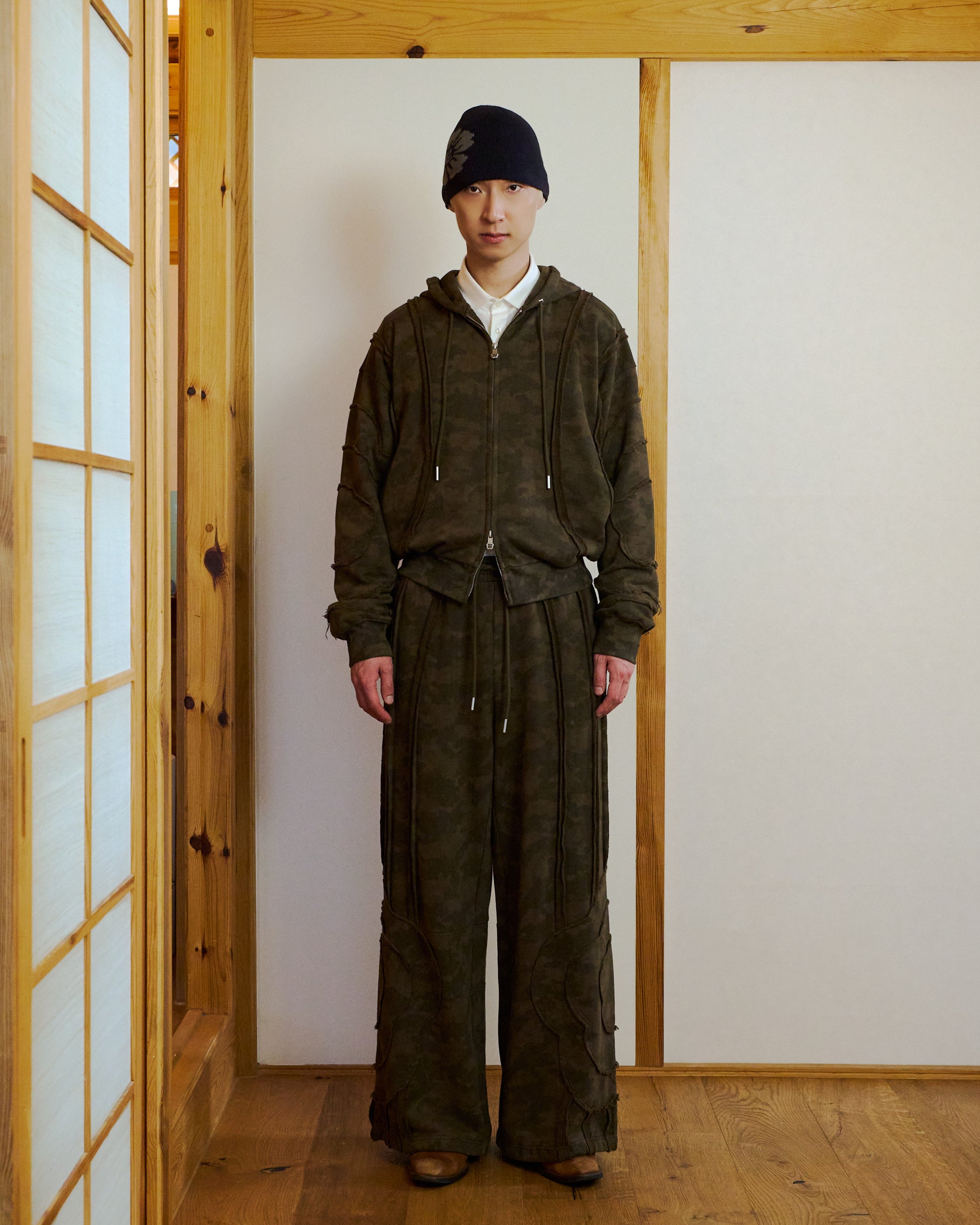 Indoors on a wooden floor, a person wears a dark camo hoodie, brown shoes, a dark beanie, and Sundae School CAMO MOCHI HAZE PANELED SWEATPANTS—wide-leg 100% cotton pants with drawstrings and raw seam panels. The space has wood paneling and white walls.