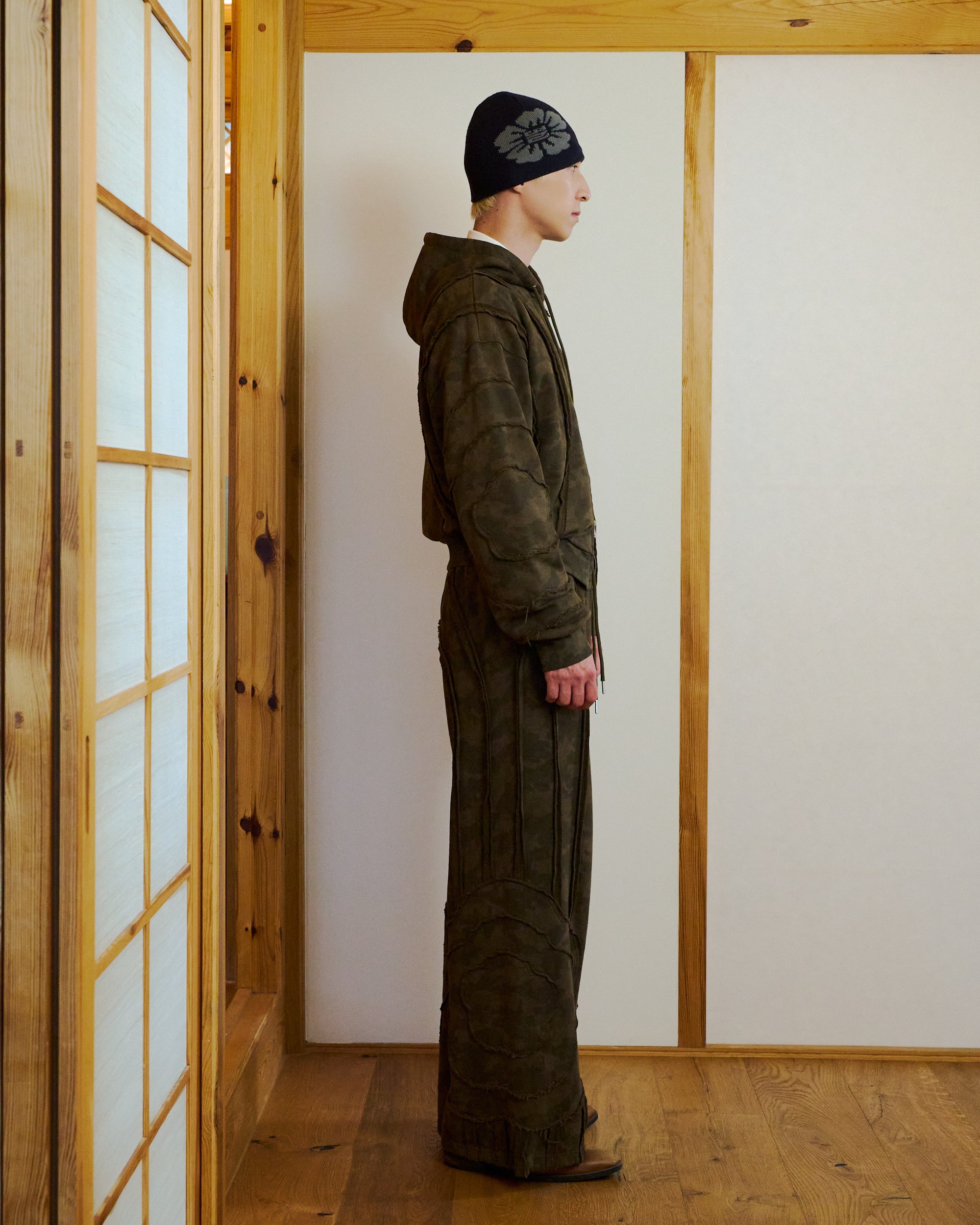 A person stands in profile in a wooden room with sliding doors, wearing Sundae School CAMO MOCHI HAZE PANELED SWEATPANTS—dark, textured, hooded 100% cotton outfit with wide pants and a black floral beanie.