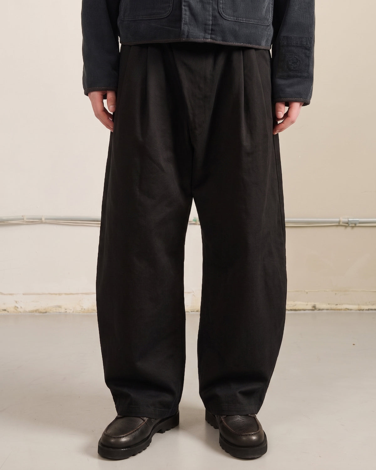 A person stands indoors wearing Sundae School's BLACK HANBOK CHINO PANTS, paired with a dark jacket and black shoes. The cropped photo focuses on the hanbok-inspired silhouette against a plain background.