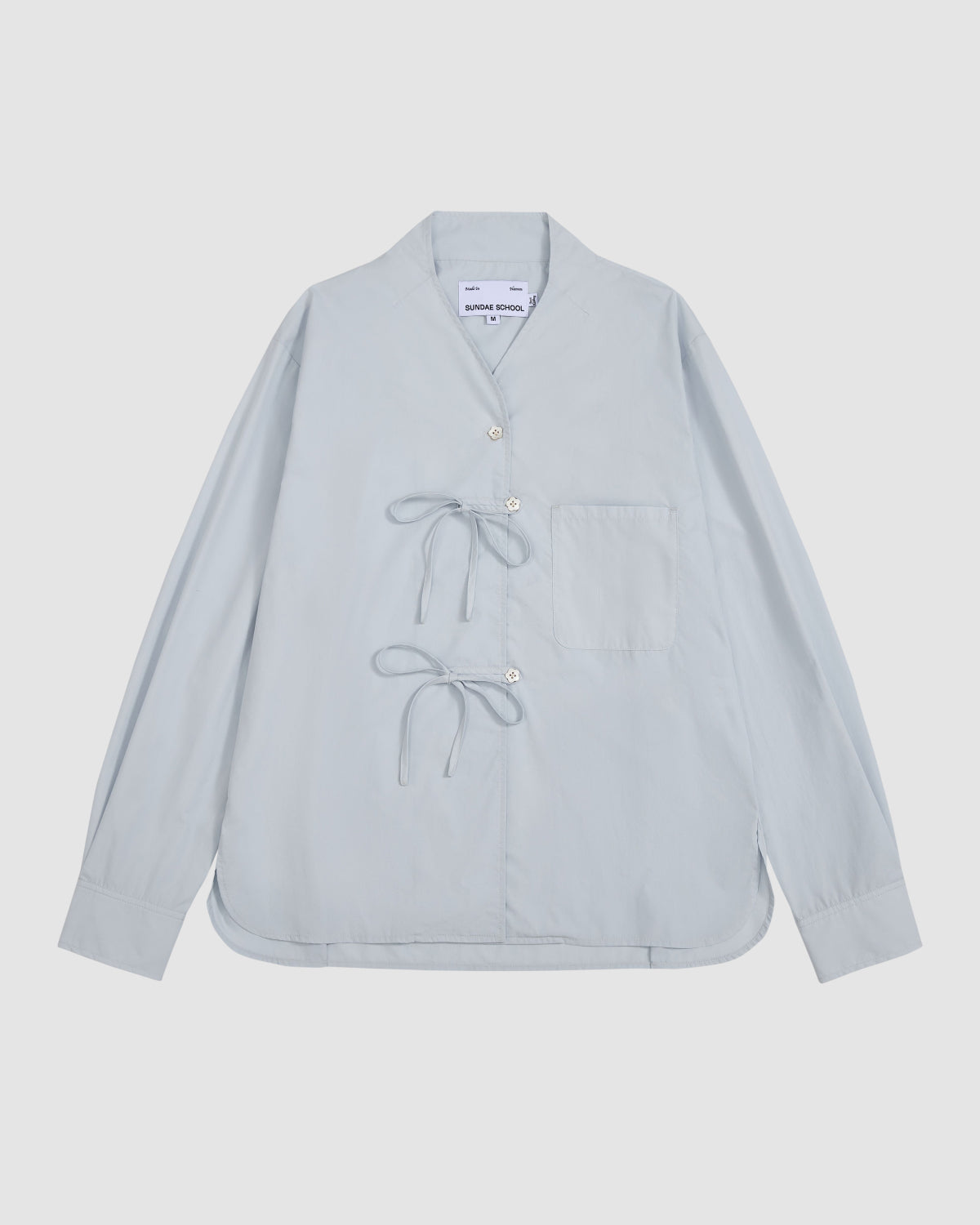 The DART COLLAR SHIRT by Sundae School is a light blue long-sleeve shirt featuring a short, round dart collar, a single chest pocket, distinctive side button closures, and two tied bows on the front. The shirt is displayed laid flat against a plain background.