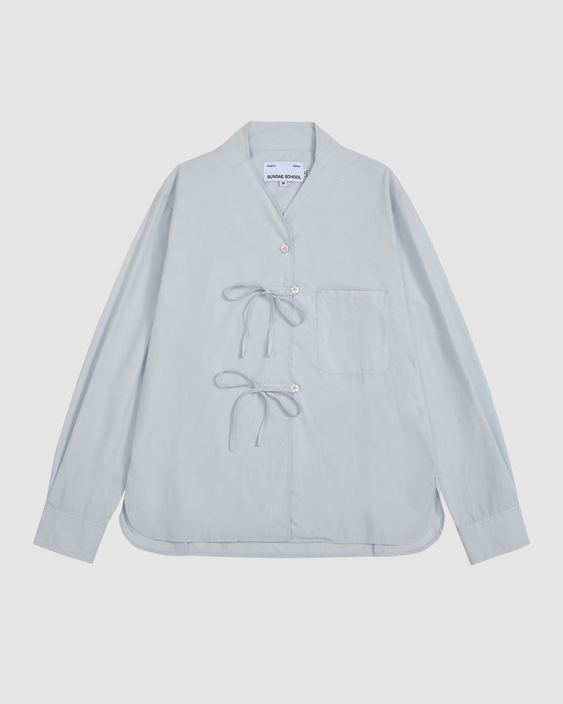 The DART COLLAR SHIRT by Sundae School is a light blue long-sleeve shirt featuring a short, round dart collar, a single chest pocket, distinctive side button closures, and two tied bows on the front. The shirt is displayed laid flat against a plain background.