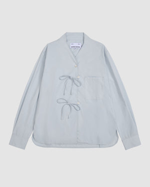 The DART COLLAR SHIRT by Sundae School is a light blue long-sleeve shirt featuring a short, round dart collar, a single chest pocket, distinctive side button closures, and two tied bows on the front. The shirt is displayed laid flat against a plain background.