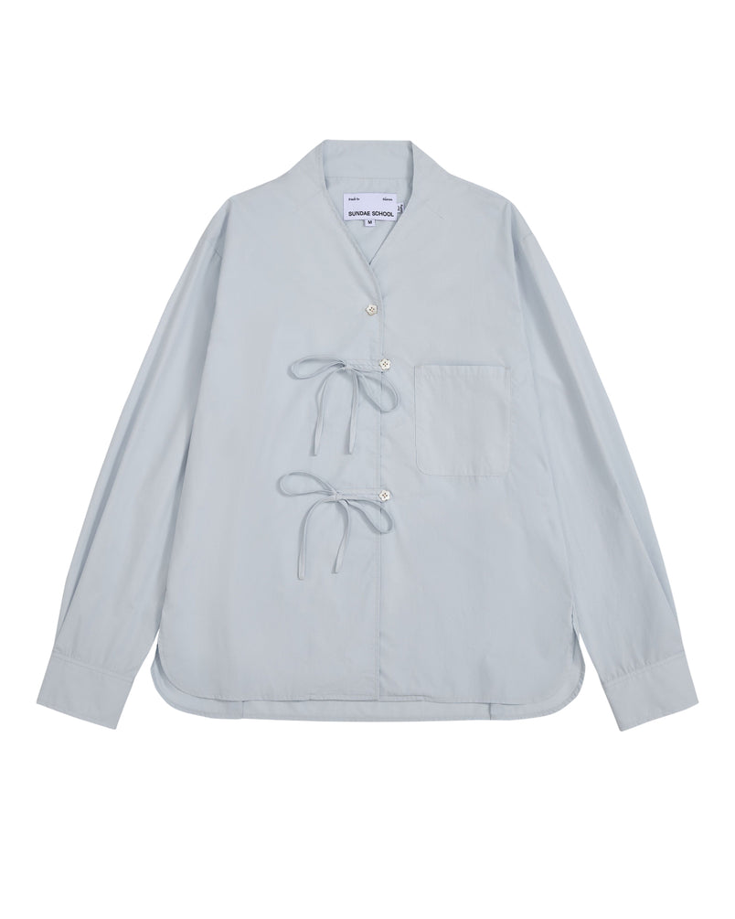 The DART COLLAR SHIRT by Sundae School is a light blue long-sleeve shirt crafted from a soft cotton-nylon blend, featuring two front tie closures, a single chest pocket, and a collarless neckline. Inspired by the relaxed silhouette of a shawl collar shirt, this men’s button-down is displayed flat against a white background.
